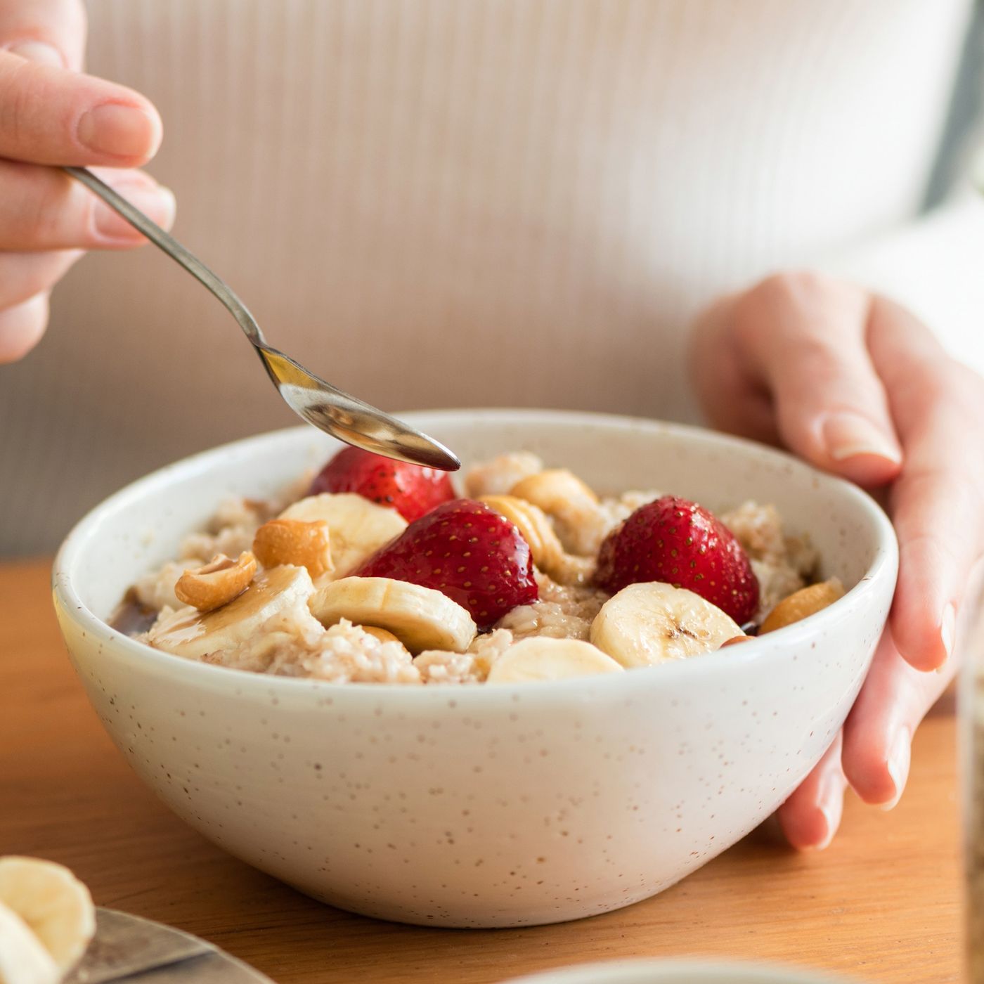 Woman eating oatmeal porridge with banana, strawberries and nuts. Healthy breakfast at the sunny morning kitchen table